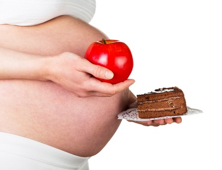 Pregnant woman holding an apple and a piece of cake isolated on white background. Healthy food concept.の写真素材