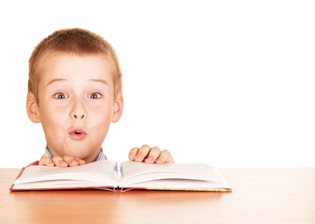 Surprised boy sitting with a book at a table isolated on white backgroundの写真素材