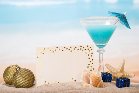 Cocktail with umbrella, blank card, Christmas balls and gifts, sea shell, starfish in the sand on the background of the sea.の写真素材