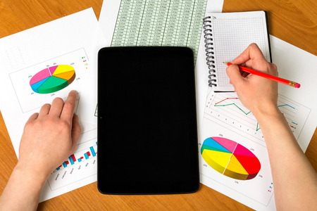 Tablet, woman's hand holding a pencil, notebook, sheets of paper account and charts on the desktop background.の写真素材
