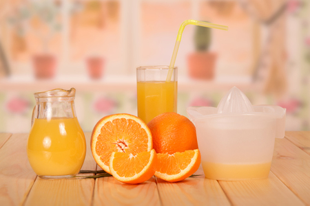 Jug and glass of fresh orange juice with a straw, orange half, the pieces and the juicer on the kitchen background.の写真素材