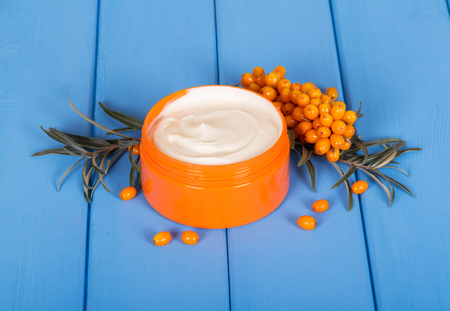 A branch with ripe sea-buckthorn berries and a jar of cream on the background of a painted tree in blue.の写真素材