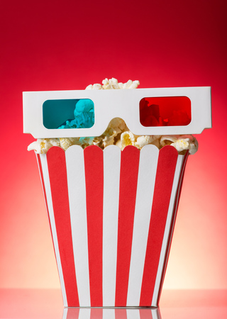 A large square box of popcorn and 3D glasses on a bright red background.