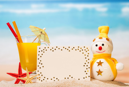 A glass of orange juice with an umbrella and tubules, a blank card, a snowman and starfish in the sand against the background of the sea.の写真素材