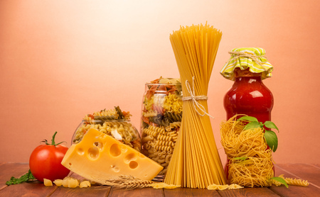 Bound spaghetti is set vertically, nest of thin vermicelli, two jars with different pasta, a bottle of ketchup, tomatoes, cheese on a red background.の写真素材