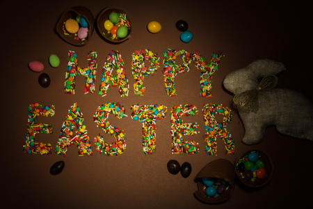 Inscription Happy Easter, lined with colorful sugar granules and confetti Easter bunny and candy on a brown background.の写真素材
