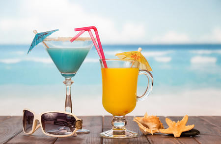 Cocktail and a glass of orange juice with umbrellas, straws, sunglasses, starfish, smooth stone and sea shell on the background of the sea.の写真素材