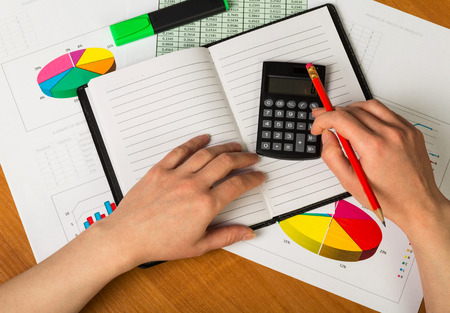Female hands holding calculator and pencil, opened the notebook, marker, sheets of paper account and charts on the desktop background.の写真素材