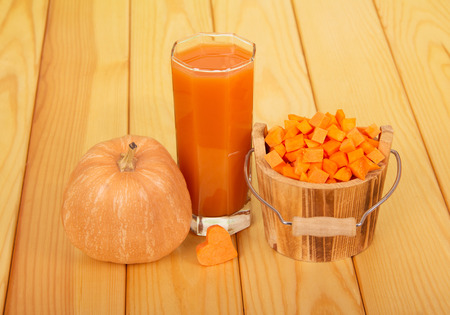 A whole ripe pumpkin, a glass of pumpkin juice, a wooden bucket with diced pumpkin cubes and a heart against a light wood background.の写真素材