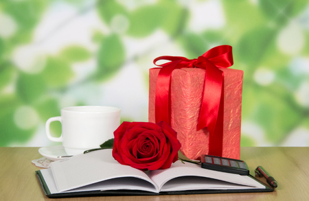 A box with a gift and ribbon, a red rose, a notebook and a pen on the tableの写真素材