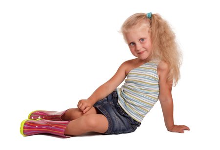 Seated charming little blond girl in a blouse, a skirt and rubber boots isolated on white background.の写真素材