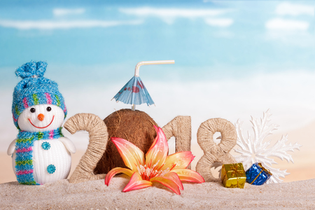 New Year inscription 2018, coconut with drinking straw and umbrella instead of the number 0, snowman and snowflake, new year gifts, flower on the sand.の写真素材