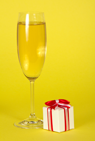A glass of sparkling wine and small souvenir decorated with ribbon on bright yellow backgroundの写真素材