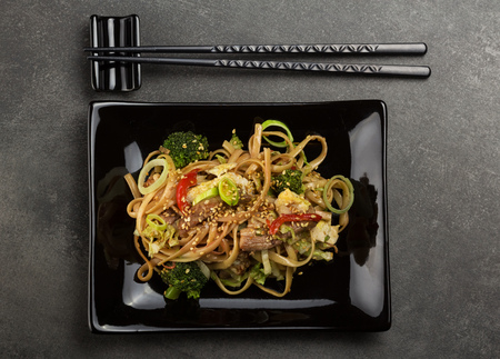 Spicy noodles with meat and vegetables in black plate, chopsticks on stand, on greyの写真素材