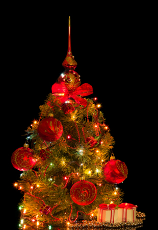 Little Christmas tree decorated with beautiful baubles and garland, nearby boxes with gifts, isolated on blackの写真素材