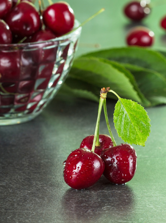 Three sweet wet berries, next to bowl of cherries, on gray surfaceの写真素材