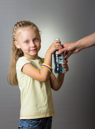 Blonde girl takes a plastic bottle with water from her mother on a gray backgroundの写真素材