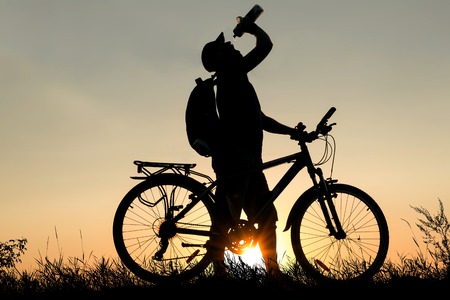 Silhouette of a man drinking water with a bicycle at sunsetの写真素材