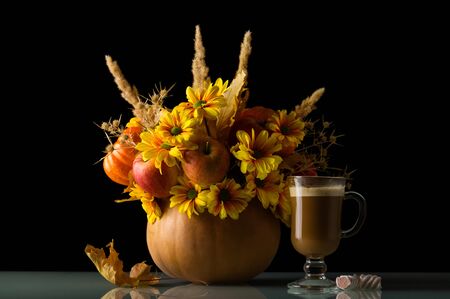 A bouquet of apples and flowers in a pumpkin, glass of cappuccino and sweets, isolated on black backgroundの写真素材