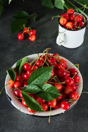 Full bowl and mug of pink cherries with leaves, on dark surfaceの写真素材