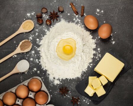 Hill of flour, broken egg on top, butter and spices in spoons on gray work surfaceの写真素材