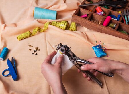 Sewing supplies, tongs for installing button on clothes in hands of seamstress, on background of beige fabricの写真素材