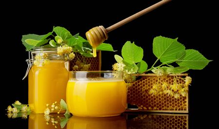 Two glass jars full of goldish honey, bee honeycombs and Linden flowers isolated on black backgroundの写真素材
