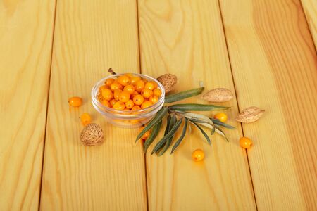 A bowl with seabuckthorn berries, a branch with leaves and almond bones next to a light wood background.の写真素材
