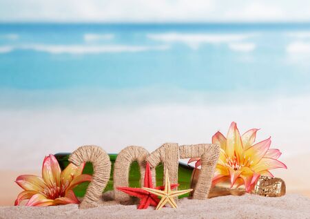 A bottle of champagne and the inscription 2017 in the sand, decorated with tropical flowers on a background of the seaの写真素材