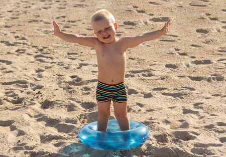 Charming smiling boy standing in lifebuoy on sandの写真素材