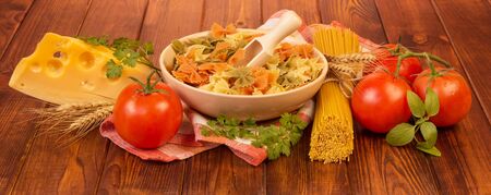 A bowl with farfalle and a wooden scoop, a bunch of spaghetti, tomatoes, cheese, parsley and basil on a dark wood background.の写真素材
