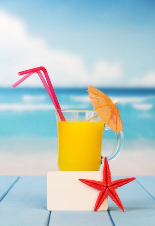 A glass of orange juice with an umbrella and straws, an empty card and a starfish against the background of the sea.の写真素材