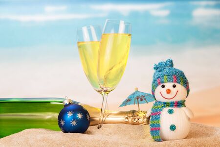 Bottle and two glasses with champagne, snowman, Christmas ball, umbrella in the sand against the sea.の写真素材