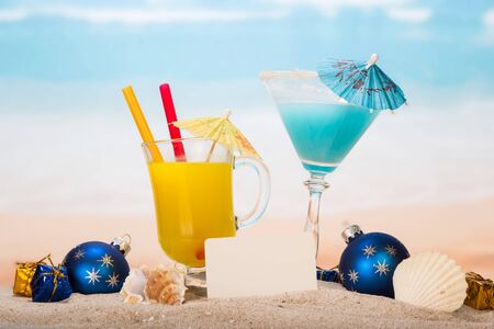 A cocktail, a glass of orange juice with umbrellas and straws, a blank card, Christmas balls and gifts, a sea shell, a shell in the sand on the background of the sea.の写真素材