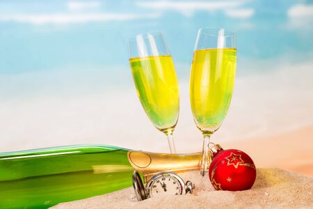 A bottle and two glasses with champagne, a clock, a Christmas ball in the sand against the background of the sea.の写真素材