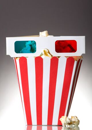 A large square box of popcorn and 3D glasses, several popcorn beside the gray background.の写真素材