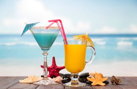 Cocktail and a glass of orange juice with umbrellas, straws, starfish, smooth stones and sea shells on the background of the sea.の写真素材
