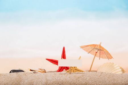 A blank card on a peg, starfish, an umbrella, a sea shell and a shell in the sand against the background of the sea.の写真素材