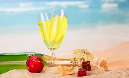 Bottle and two glasses of champagne, Christmas ball and gifts, umbrella, starfish, seashell in the sand on the sea background.の写真素材