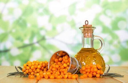 An overturned bucket of sea buckthorn berries, a branch with berries and a bottle of oil on abstract green background.の写真素材