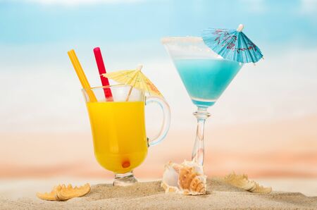 Cocktail and a glass of orange juice with umbrellas and tubes, starfish, sea shell in the sand against the background of the sea.の写真素材