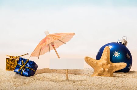 A blank card on a peg, an umbrella, a Christmas ball and gifts, a starfish in the sand against the background of the sea.の写真素材