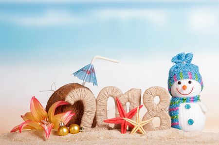 New Year inscription 2018, snowman and Christmas balls, coconut with drinking straw and umbrella, starfish, flower in the sand.の写真素材