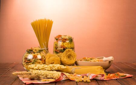 Banks with various pasta, spaghetti bunches, a bowl of farfalle and a shovel, nests of thin vermicelli and noodles on a red background.の写真素材