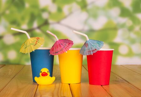 Three colored disposable Cup, with a straw and an umbrella next to a child's toy, on beige wooden tableの写真素材