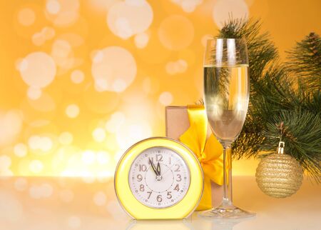 Christmas pine branch with toy, glass of champagne, gift and clock on bright yellow backgroundの写真素材
