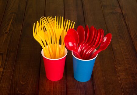 Set of bright disposable spoons and forks in plastic cups, on brown background of wooden tableの写真素材