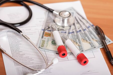 Phonendoscope, test tubes for blood testing, results of survey and money on background of tableの写真素材