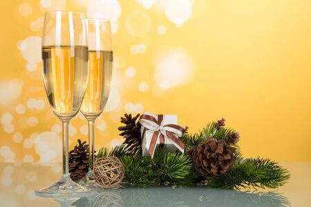 New Year pine branch with cones, gift and wine glasses with champagne, on light yellow backgroundの写真素材