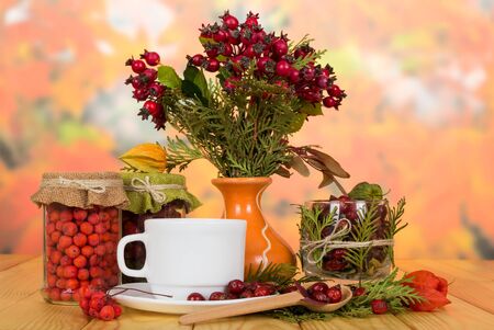 Berries of mountain ash for tea, vase with bouquet of wild rose, cup and spoon on beige wooden tableの写真素材